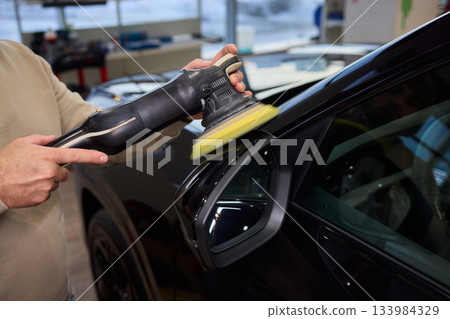 Polishing black car surface thoroughly, Detailed buffing to achieve showroom quality shine, Expert technician uses orbital buffer and foam pad to enhance vehicle gloss 133984329