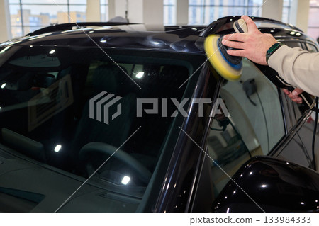 Polishing black car surface thoroughly, Detailed buffing to achieve showroom quality shine, Expert technician uses orbital buffer and foam pad to enhance vehicle gloss 133984333
