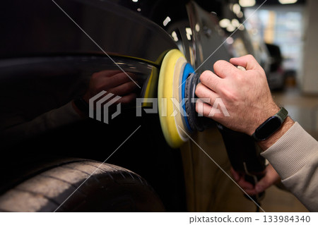 Polishing black car surface thoroughly, Detailed buffing to achieve showroom quality shine, Expert technician uses orbital buffer and foam pad to enhance vehicle gloss 133984340