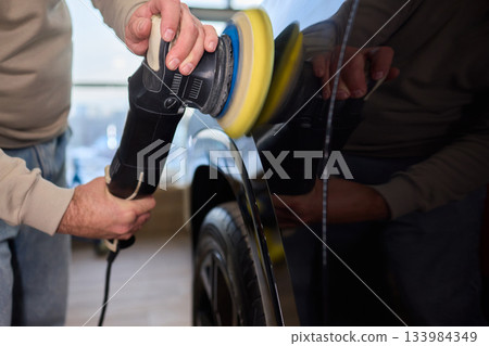 Polishing black car surface thoroughly, Detailed buffing to achieve showroom quality shine, Expert technician uses orbital buffer and foam pad to enhance vehicle gloss 133984349
