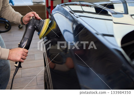 Polishing black car surface thoroughly, Detailed buffing to achieve showroom quality shine, Expert technician uses orbital buffer and foam pad to enhance vehicle gloss 133984350