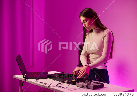 Female DJ mixing on turntables, colorful lighting studio, focused expression, hands adjusting mixer and laptop controls, wearing headphones, modern electronic music vibe, cinematic lighting 133984376