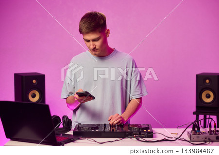Young DJ with headphones, Skilled DJ engaging with equipment in lively environment, Energetic DJ prepping for live performance amid colorful lighting and fashionable attire 133984487