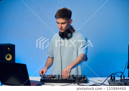Young DJ with headphones, Skilled DJ engaging with equipment in lively environment, Energetic DJ prepping for live performance amid colorful lighting and fashionable attire 133984488