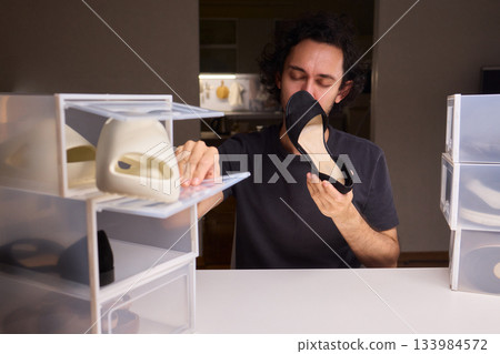 Man sniffing black shoes from plastic box, checking odor condition in dim storage room with stacked clear drawers and fluorescent lamp 133984572
