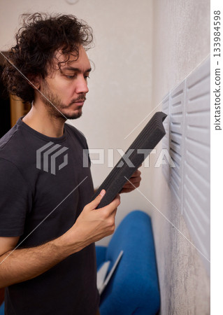 Closeup of foam insulation method, Hand pressing textured acoustic foam onto wall surface, Detailed shot of fingers securing black foam panels on gray wall Comparison of different foam rubber and 133984598