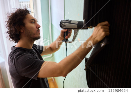 Craftsman carefully detaching acoustic panels indoors, Individual skillfully peels acoustic insulation from wall using heat and precise hand movements for studio setup 133984634