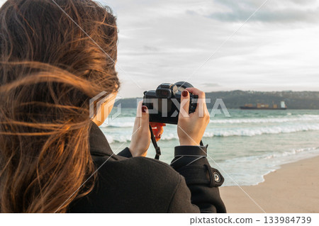 Smiling female photographer holding a camera on a sunny beach, enjoying golden hour light and capturing outdoor moments on the coastline. Smiling female photographer holding a camera on a sunny beach, enjoying golden hour light and capturing outdoor moments on the coastline. 133984739