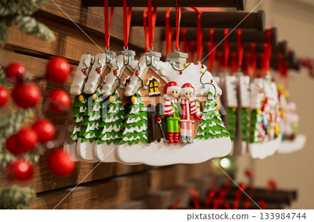 Row of Christmas house ornaments hanging on red ribbons at festive market stall, holiday decoration background with warm lights and wood texture. 133984744