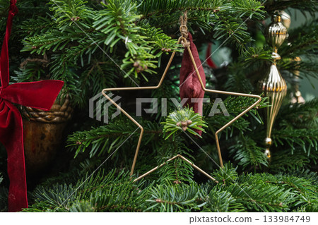 Minimalist golden star ornament hanging on fir branch. Modern christmas decor with clean lines and natural evergreen texture. Minimalist golden star ornament hanging on fir branch. Modern christmas decor with clean lines and natural evergreen texture. 133984749