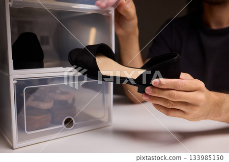 Man sniffing black shoes from plastic box, checking odor condition in dim storage room with stacked clear drawers and fluorescent lamp Man sniffing black shoes from plastic box, checking odor condition in dim storage room with stacked clear drawers and fluorescent lamp 133985150