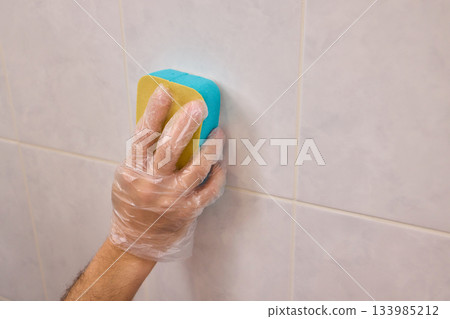 A Man Diligently Cleaning a Tiled Bathroom Wall with His Gloves and a Scrubber Tool A Man Diligently Cleaning a Tiled Bathroom Wall with His Gloves and a Scrubber Tool 133985212