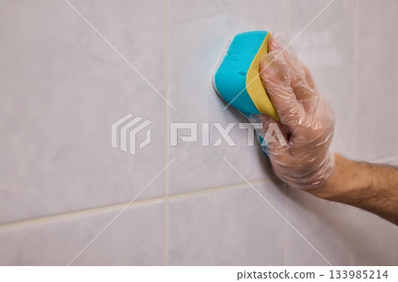 A Man Diligently Cleaning a Tiled Bathroom Wall with His Gloves and a Scrubber Tool 133985214