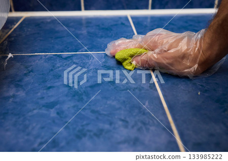 Hand cleaning grout, Scrubbing ceramic tile joints professionally, Detailed hand scrub of grout with sponge and gloves, Closeup of hand using sponge to clean grout lines Hand cleaning grout, Scrubbing ceramic tile joints professionally, Detailed hand scrub of grout with sponge and gloves, Closeup of hand using sponge to clean grout lines 133985222