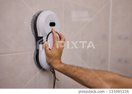 Smart robot performing quick hygienic maintenance in bathroom space, Modern robotic cleaner diligently scrubbing grout lines adjacent to toilet in bright tiled room 133985226