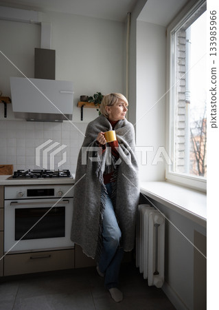 Middle aged woman staying warm in cold unheated apartment wrapping in plaid and drinking hot tea. 133985966