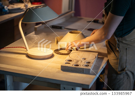 Man carpenter manufacturing wooden lamp in carpentry workshop, choosing wire for furniture object. Man carpenter manufacturing wooden lamp in carpentry workshop, choosing wire for furniture object. 133985967