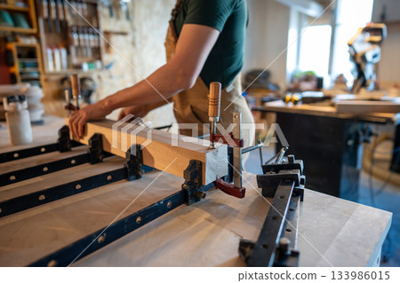 Man carpenter working in carpentry workshop, compressing glued wooden blocks at table with clamps. 133986015