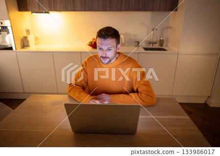 Adult businessman freelancer works at home online on laptop, sitting in kitchen late at night. 133986017