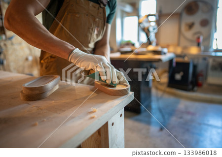 Man carpenter sanding wooden coaster for water glasses, working process in small carpentry workshop. 133986018