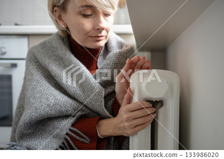 Tired middle aged woman freezing in plaid and warming hands on central heating radiator in apartment 133986020