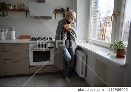 Middle aged woman staying warm in cold unheated apartment wrapping in plaid and drinking hot tea. 133986023