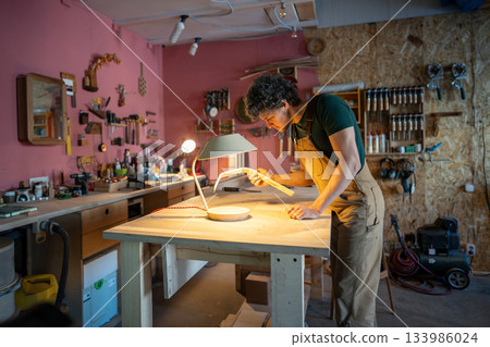 Man carpenter working on lamp in carpentry workshop, precisely evaluating quality of wooden part. 133986024