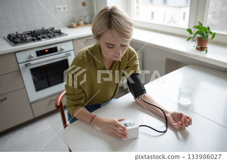Middle aged woman sits at table measuring blood pressure with tonometer, seasonal health problems. 133986027