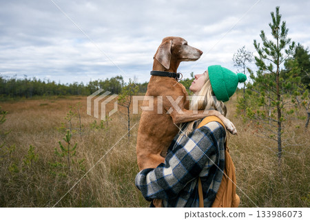 Affectionate woman hiker embracing Magyar Vizsla in autumnal field, backpack adventure in fall 133986073
