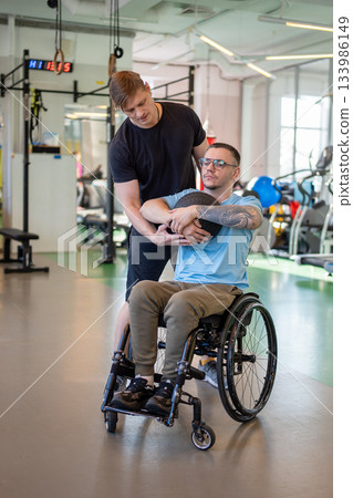 Personal coach supporting disabled man in wheelchair during power workout with medicine ball in gym. Personal coach supporting disabled man in wheelchair during power workout with medicine ball in gym. 133986149