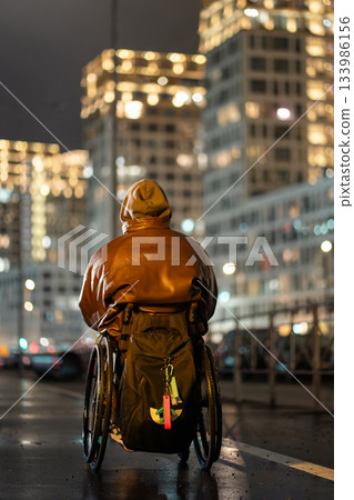 Man in wheelchair in hood with backpack walking alone late at night under color rgb light in city. 133986156