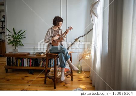 Young woman playing ukulele alone by window at home, repeating chord progressions with focus 133986164