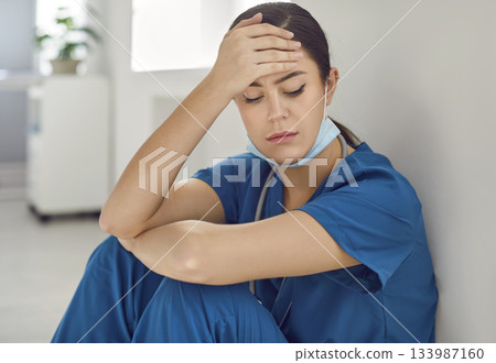 Exhausted, stressed, sad woman doctor sitting on floor by wall after long day at work Exhausted, stressed, sad woman doctor sitting on floor by wall after long day at work 133987160