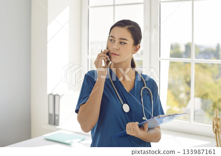 Doctor or nurse talking by mobile phone standing with clipboard notepad in medical clinic. Doctor or nurse talking by mobile phone standing with clipboard notepad in medical clinic. 133987161