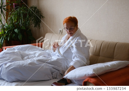 old woman in robe sits on bed in bedroom, takes out glasses, reads book, relaxes in morning. Senior woman sitting pensively Vintage, cozy, relaxed, contemplative, natural light, serenity, elegant old woman in robe sits on bed in bedroom, takes out glasses, reads book, relaxes in morning. Senior woman sitting pensively Vintage, cozy, relaxed, contemplative, natural light, serenity, elegant 133987234