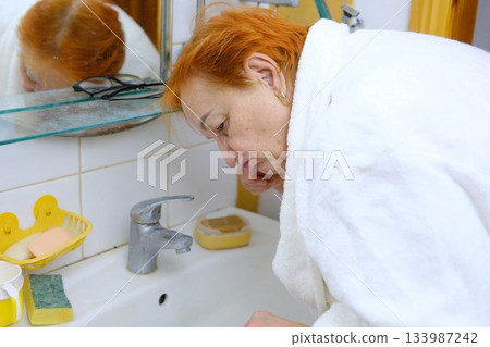 red-haired woman in a robe elderly senior woman is cleaning brushing his teeth in front of mirror in bathroom. Dental hygiene, vitality and beauty concepts 133987242