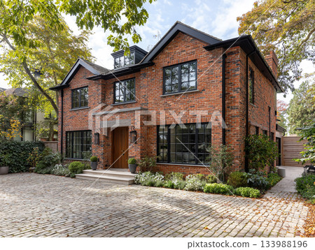 A modern luxury house featuring dark red brick, black-framed windows, a slate roof, and a large integrated garage, surrounded by lush mature trees and a paved driveway. 133988196