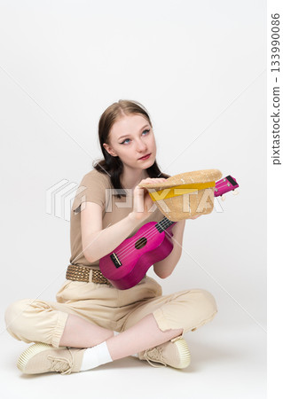Female musician with ukulele holds straw hat in front of her for donations and tips, sitting cross legged on floor, looking up. Woman ukulele artist Caucasian ethnicity in casual, comfortable outfit 133990086