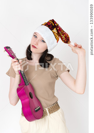 Young female musician attired in Santa Claus hat and she holds ukulele in one hand and pom pom in other. Her facial expression is characterized by playful and friendly demeanor, as she looks at camera 133990089