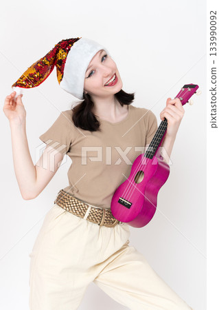Young female musician, attired in red Santa hat and holding ukulele, poses playfully, directs gaze toward camera. Beautiful woman engages in flirtatious behavior, exhibiting smile, protruding tongue Young female musician, attired in red Santa hat and holding ukulele, poses playfully, directs gaze toward camera. Beautiful woman engages in flirtatious behavior, exhibiting smile, protruding tongue 133990092
