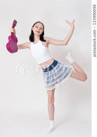 Extraordinary woman holding ukulele and doing flexibility exercises, dancing and smiling as she looks camera. Female ukulele player wearing tank top, short skirt, socks. She looks happy. Full length Extraordinary woman holding ukulele and doing flexibility exercises, dancing and smiling as she looks camera. Female ukulele player wearing tank top, short skirt, socks. She looks happy. Full length 133990098