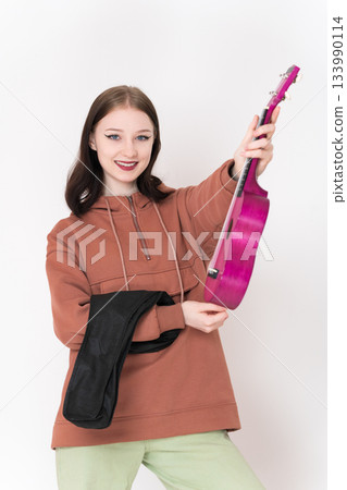 Positive young woman ukulelist holds ukulele in her hands and smiles, looking at camera. 18 year old woman is wearing brown hoodie and light green jeans Positive young woman ukulelist holds ukulele in her hands and smiles, looking at camera. 18 year old woman is wearing brown hoodie and light green jeans 133990114