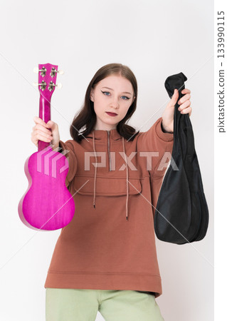 Ukulele artist holds ukulele in one hand, instrument case in other, and extends both arms forward in front. Beautiful woman in casual clothing with brown hoodie and light green jeans Ukulele artist holds ukulele in one hand, instrument case in other, and extends both arms forward in front. Beautiful woman in casual clothing with brown hoodie and light green jeans 133990115