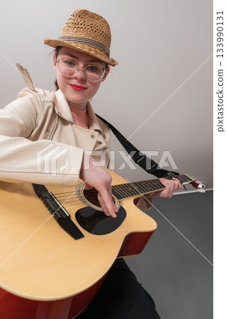 Cheerful female guitarist with glasses and straw hat practicing acoustic guitar, looking at camera. Low angle and dynamic perspective makes performance look lively atmosphere and full of energy Cheerful female guitarist with glasses and straw hat practicing acoustic guitar, looking at camera. Low angle and dynamic perspective makes performance look lively atmosphere and full of energy 133990131