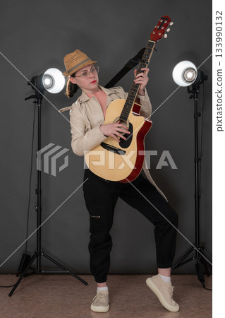Young woman guitarist strumming six string acoustic classical guitar, standing full length on stage. Serious woman musician dressed in beige leather jacket, straw hat, black jeans pants and sneakers Young woman guitarist strumming six string acoustic classical guitar, standing full length on stage. Serious woman musician dressed in beige leather jacket, straw hat, black jeans pants and sneakers 133990132