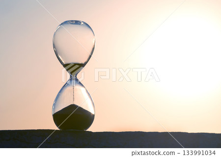 Hourglass framed by a radiant sunset, sand trickling down in soft light Hourglass framed by a radiant sunset, sand trickling down in soft light 133991034