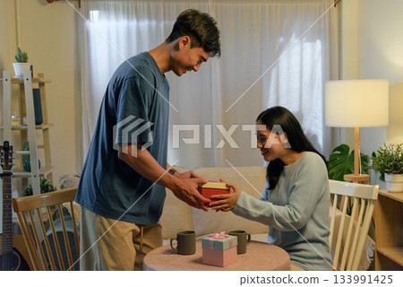 Happy young couple in love, man gives woman a special red gift box for romantic valentine day celebration at home, sharing affection and joy indoors with lovely atmosphere. Happy young couple in love, man gives woman a special red gift box for romantic valentine day celebration at home, sharing affection and joy indoors with lovely atmosphere. 133991425
