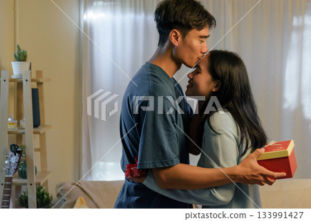 Young happy couple in love, woman man at home, share romantic valentine day affection, man kisses woman forehead, celebrating special moment with gifts. 133991427