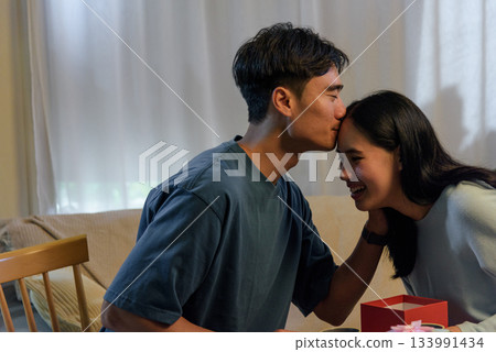 Young happy couple shares sweet love at home, man kissing woman forehead, romantic valentine day celebration with surprise gift, expressing deep affection, tender special moments together. 133991434