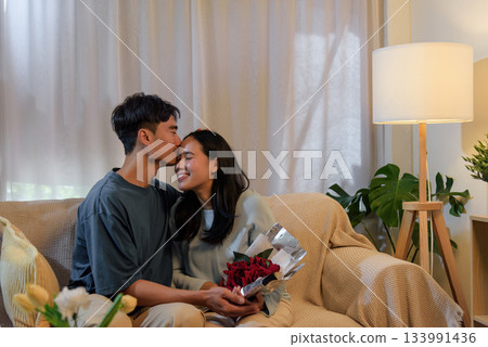 Young couple happy love, man woman share romantic valentine day at home, man kisses woman on forehead while she holds beautiful red roses, perfect moment expressing affection. 133991436